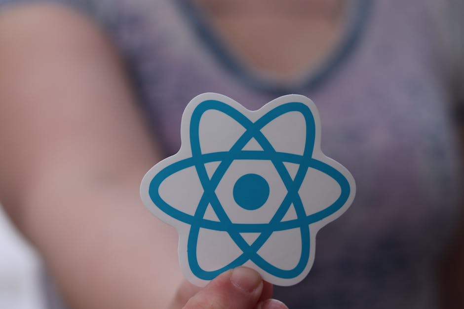 A person holding a cutout paper with a blue atomic symbol, featuring a central circle and several intersecting elliptical orbits, in front of a blurred background that suggests an indoor setting with 